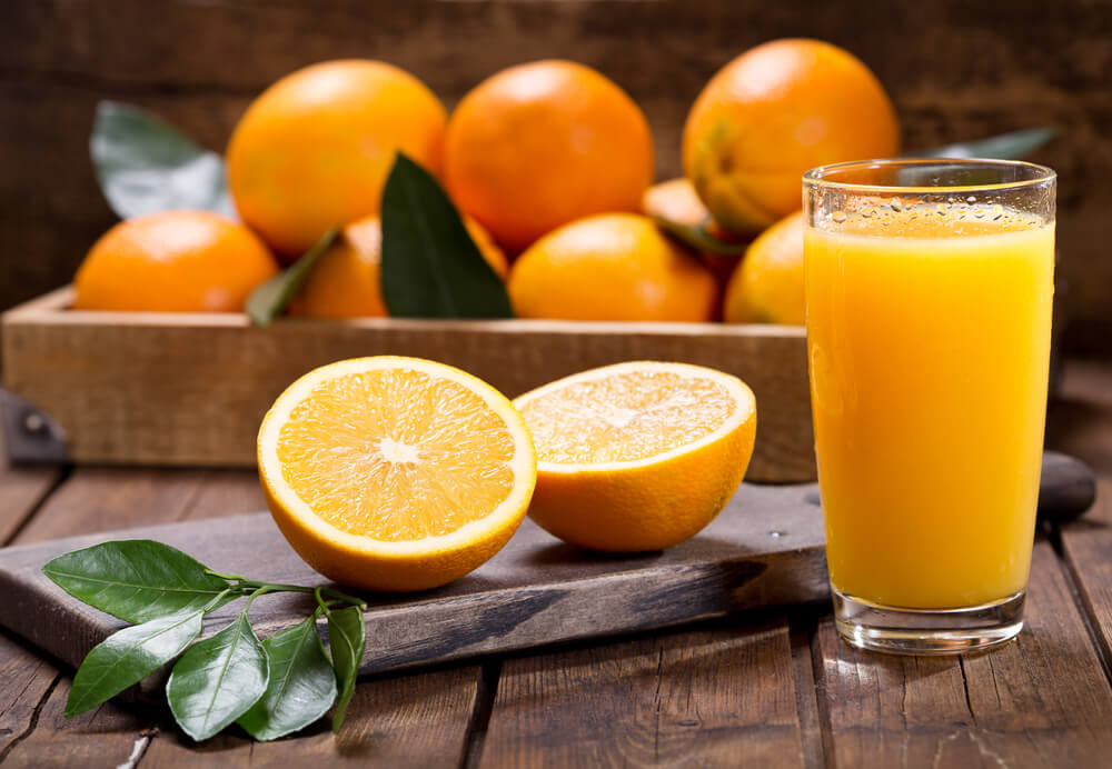 Свежевыжатый натуральный апельсиновый сок в стакане. Рядом с ним на деревянной доске лежат два апельсина. 