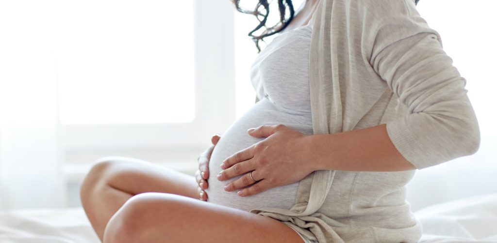 Die schwangere Frau sitzt auf dem Bett und berührt sanft ihren Bauch.