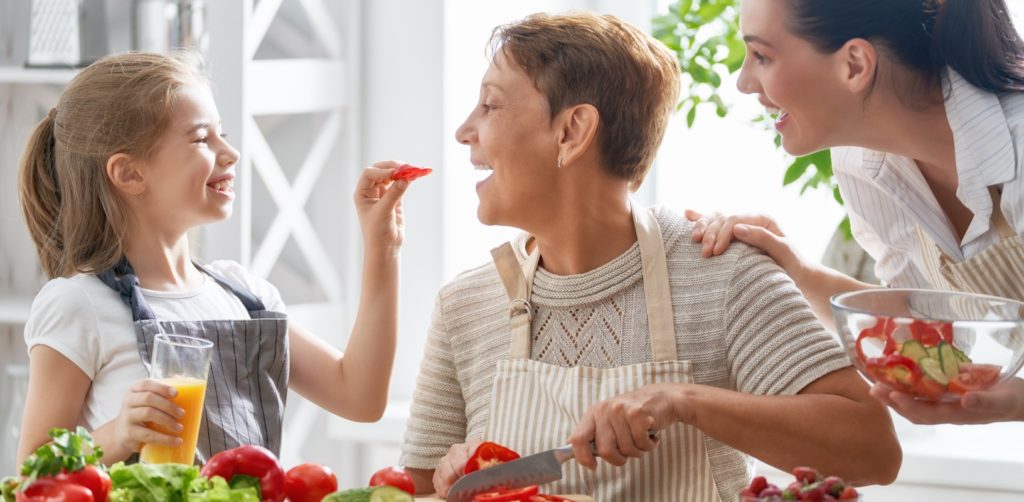 A caring granddaughter is feeding her grandma with refreshing vegetables. She is preparing summer lunch together with another granddaughter.