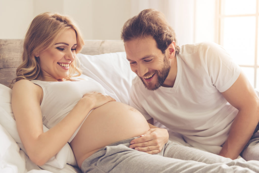 Eine schöne schwangere Frau und ihr Partner verbringen Zeit miteinander im Bett, wo er begeistert ihren dicken Bauch betrachtet.