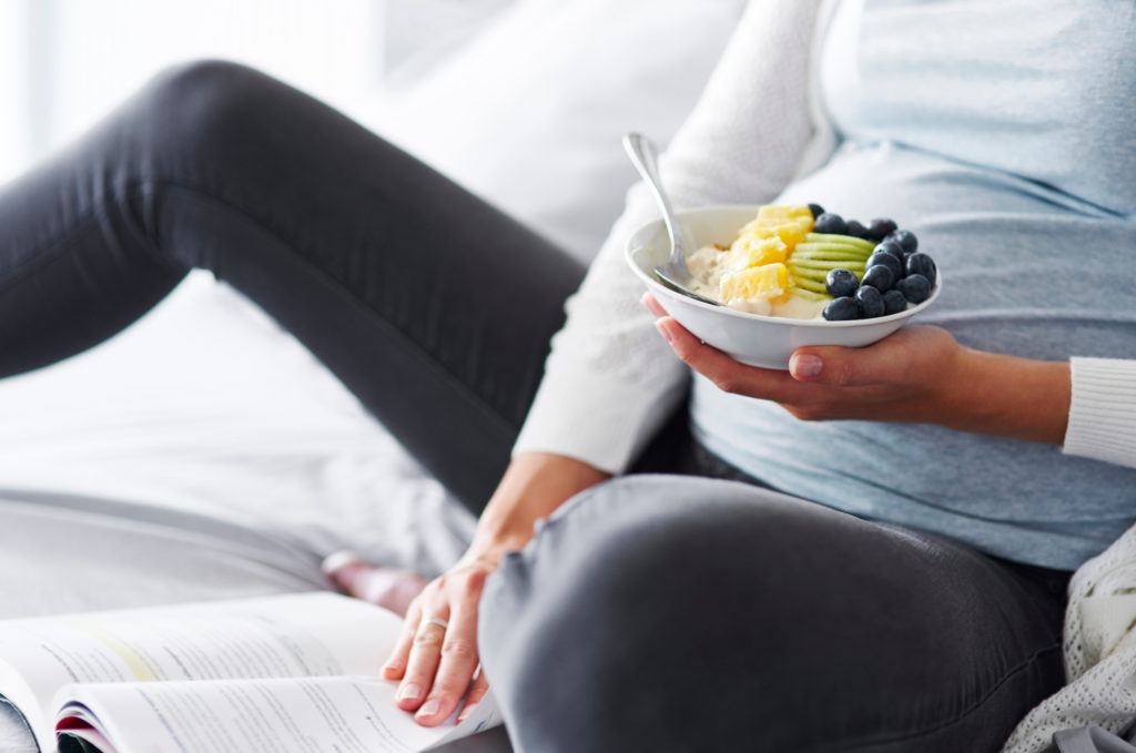 Беременная женщина лежит в постели. Она ест здоровый завтрак и читает книгу.