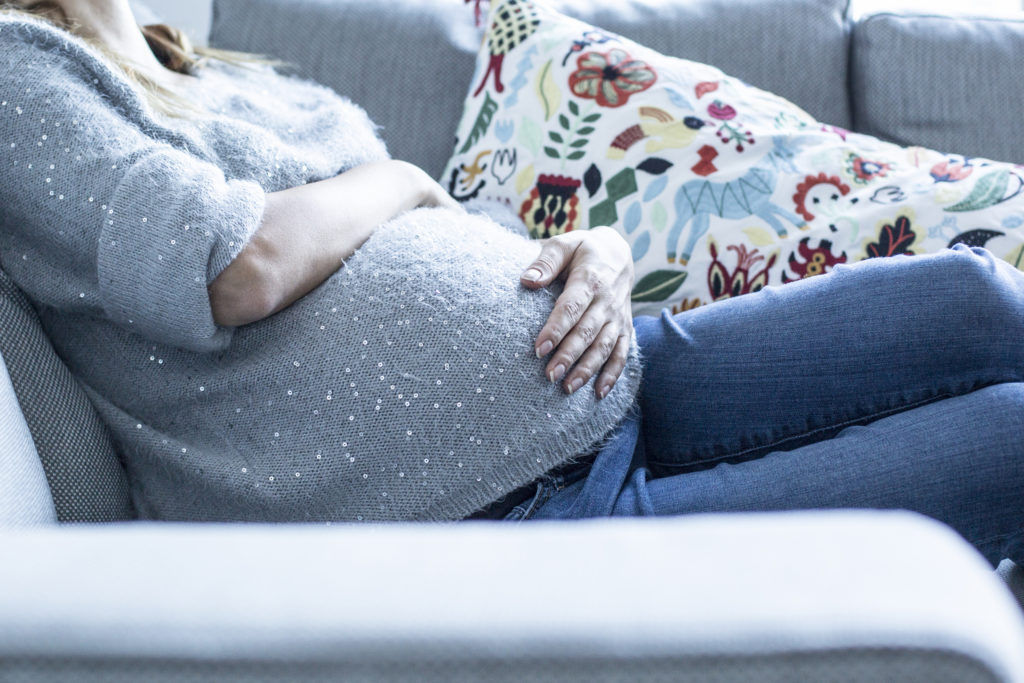 Молодая беременная женщина сидит на диване и гладит живот. 