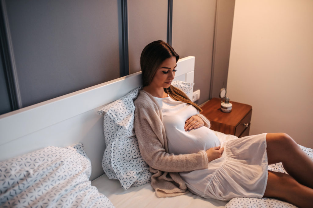 Беременная женщина сидит на кровати, смотрит и гладит живот. 