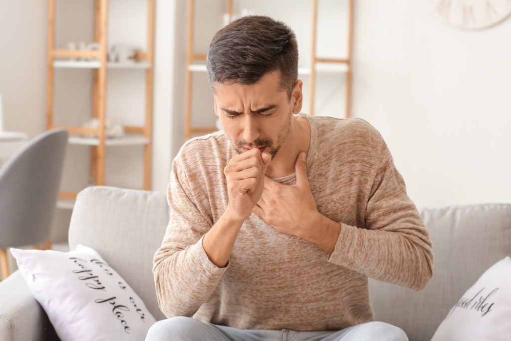 A man sitting on the sofa and coughing because he is suffering from heartburn.