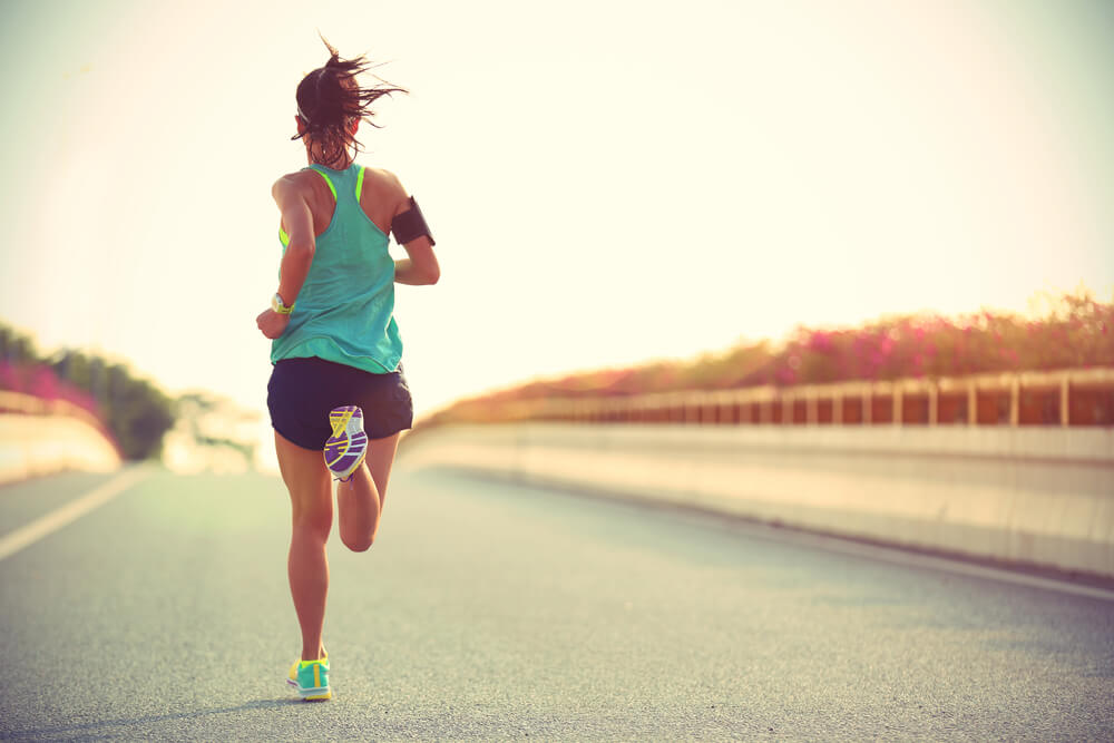 Fit ženska v športnih oblačilih teče po asfaltni cesti proti sončnemu zahodu.