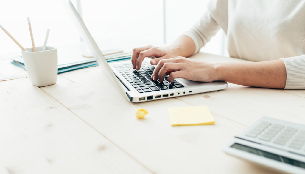 Eine Frau in einem weißen Pullover sitzt an einem Tisch und schreibt auf einem Laptop.