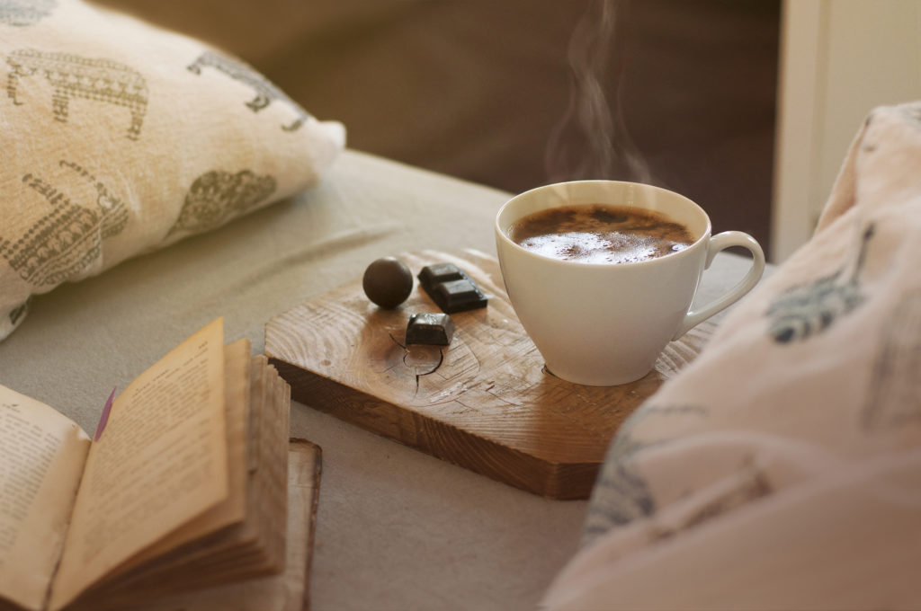 Freshly made cup of coffee and pieces of chocolate on a wooden platter placed on bed.