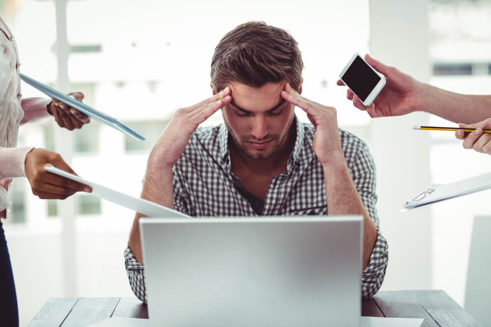 Muškarac na poslu sjedi pred računalom i drži se za glavu, jer je pod velikim stresom, budući da ga pritiskaju sa svih strana.