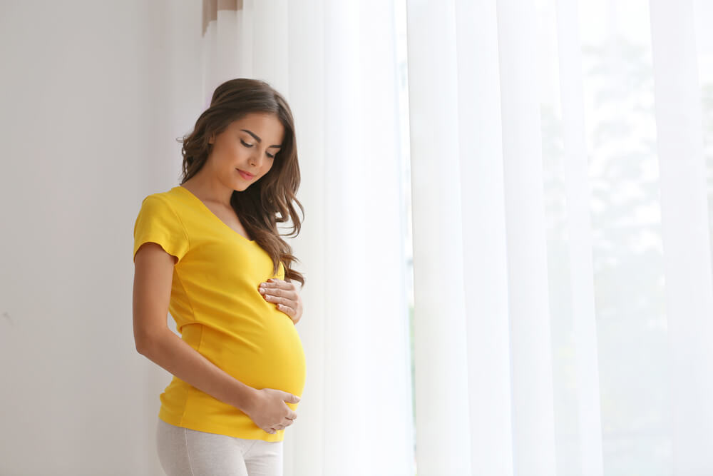 Eine junge schwangere Frau in einem gelben Shirt steht zu Hause am Fenster und streichelt ihren Bauch.