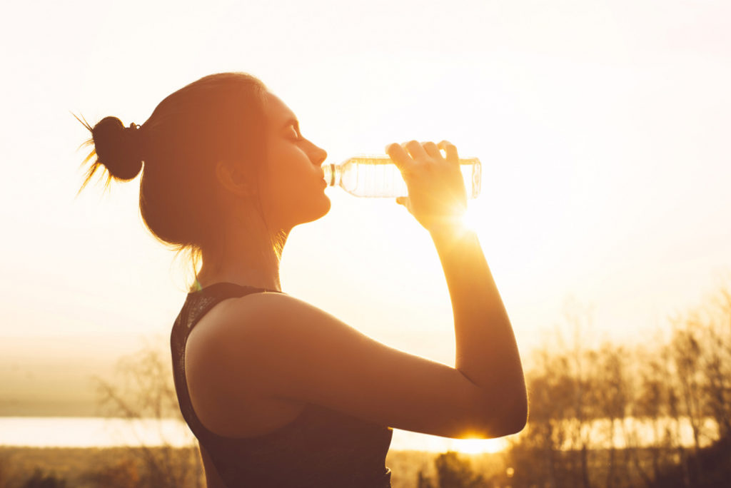 A sportswoman is getting hydrated with a bottle of water during her workout in the summer sunset.