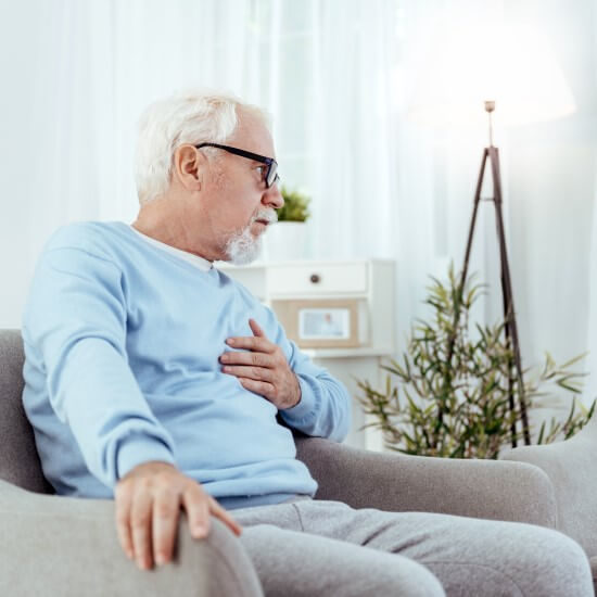 An older man is sitting on the sofa and clutching at his chest because he is suffering from heartburn.