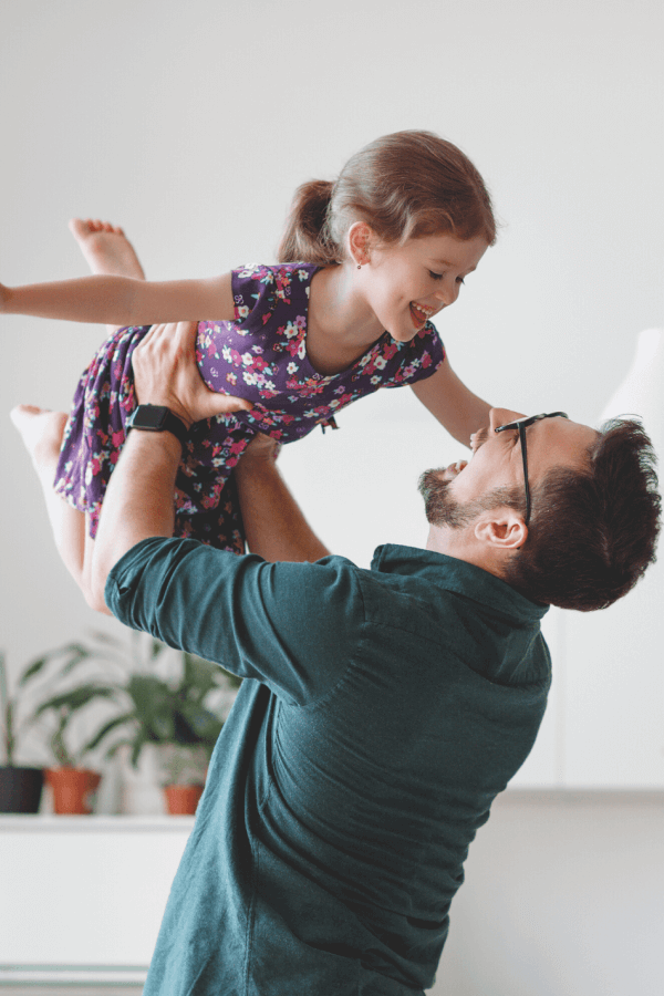 A happy dad is lifting his daughter high up in the air. They are both laughing hard.