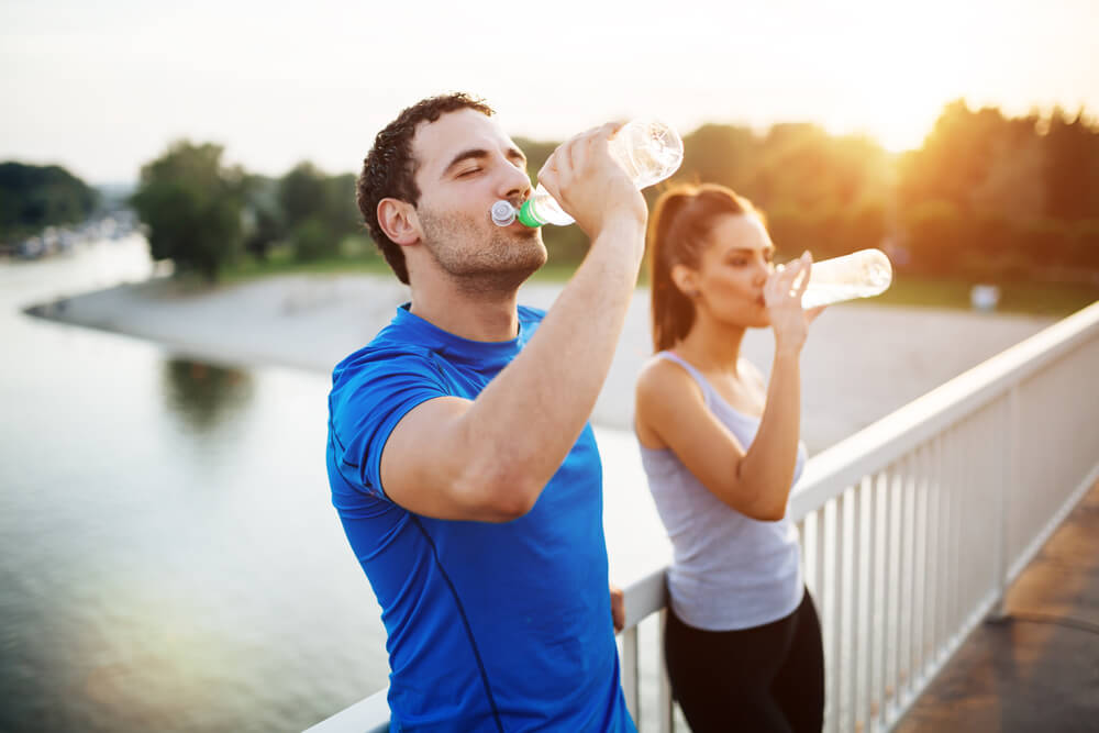 A man and a woman are standing on a bridge and drinking water from a plastic bottle because they have just finished their jog.