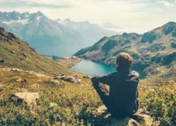 A traveler who is meditating by relaxing on a rock and admiring the magnificent view of the mountains and the lake.