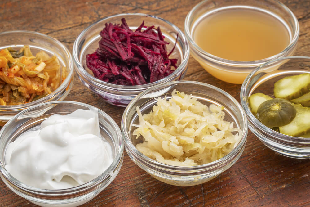 Sechs Glasbehälter mit fermentierten Lebensmitteln, die geschnittene Rüben, Apfelessig, Kokosmilchjoghurt und geschnittene Gurken und Sauerkraut enthalten.