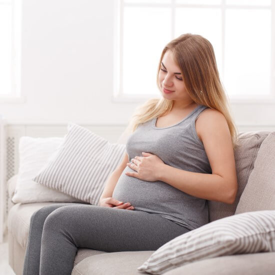 A woman wearing grey clothes is sitting at home on the sofa and caressing her stomach with both her hands.