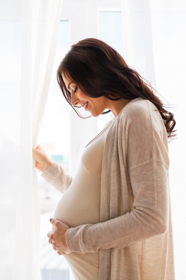 A happy young pregnant woman with a big stomach is standing by the window and caressing her stomach.