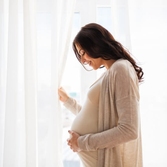 A happy young pregnant woman with a big stomach is standing by the window and caressing her stomach.