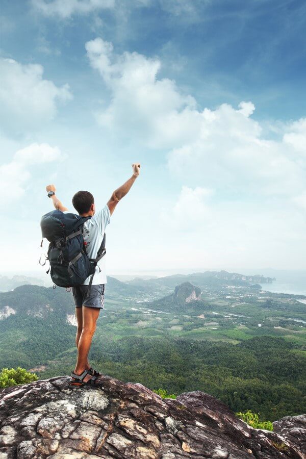 A young traveler is standing at the mountain top and holding his hands high up in the air to express happiness for conquering the peak.