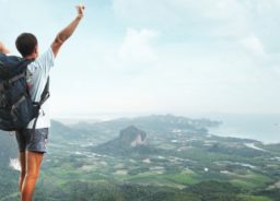 A young traveler is standing at the mountain top and holding his hands high up in the air to express happiness for conquering the peak.