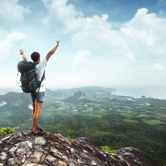 A young traveler is standing at the mountain top and holding his hands high up in the air to express happiness for conquering the peak.