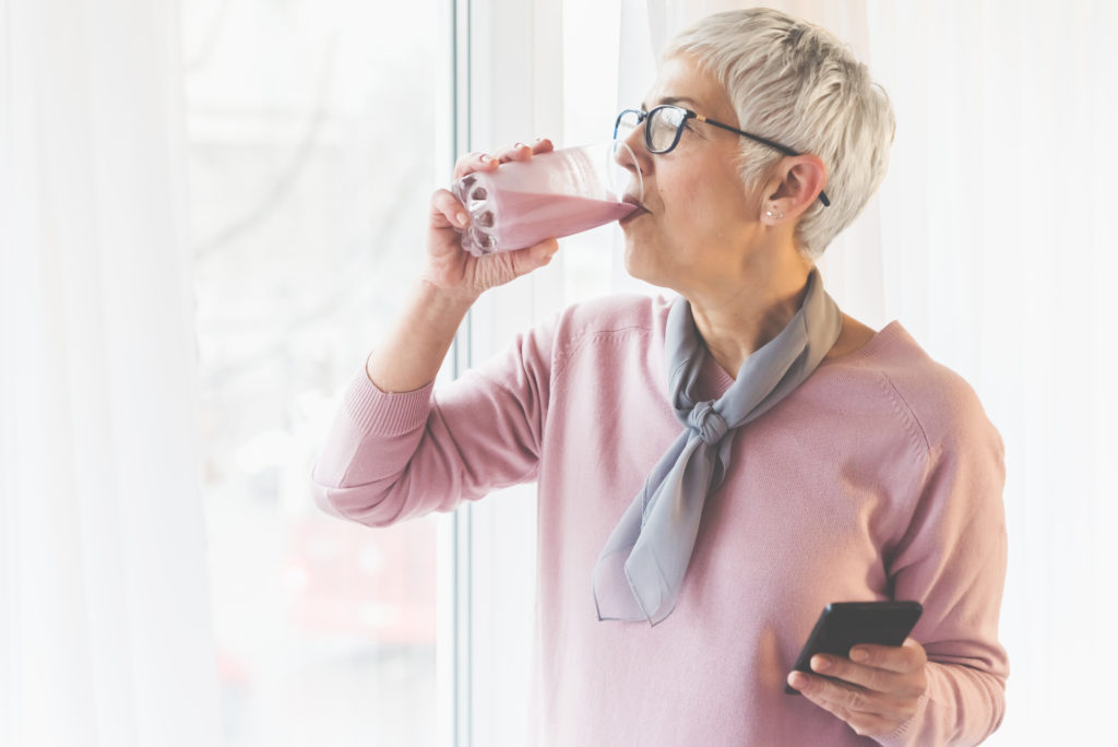 Eine ältere Frau hält in einer Hand einen leckeren und gesunden Smoothie und trinkt es und mit der anderen Hand hält sie ein Telefon.