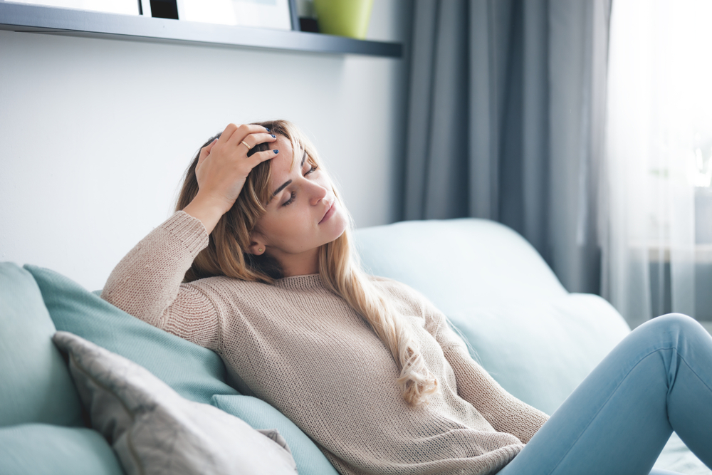 A tired young woman is sitting on a light blue sofa with her eyes closed and holding her head.