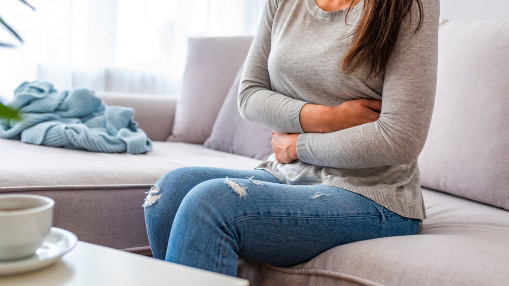 Eine junge Frau mit Schmerzen sitzt zu Hause auf der Couch und hält sich am Bauch.