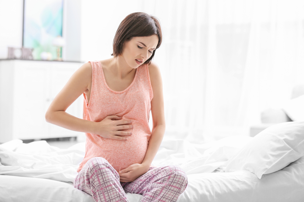 Eine schwangere Frau mit Schmerzen sitzt auf dem Bett und hält sich an ihrem Bauch fest.