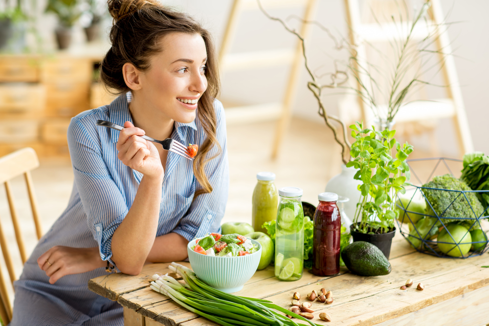 Eine junge fröhliche Frau isst einen Salat mit gesunden Zutaten und sitzt an einem Holztisch voller grünem Gemüse, Obst und frisch gepressten Säften.