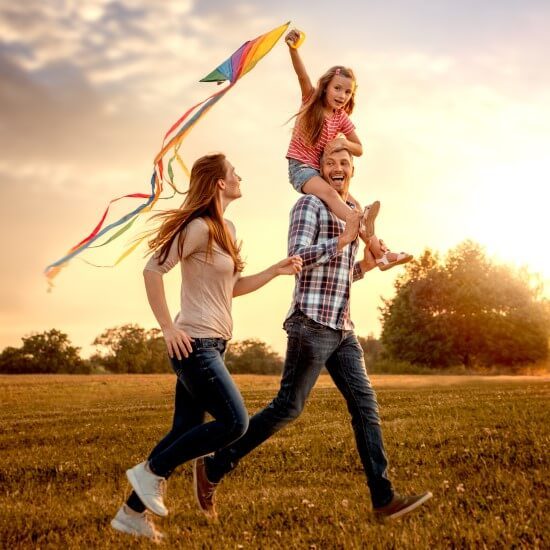 A happy family – father, mother and daughter sitting on her father’s shoulders – they are flying a kite on the meadow while the sun is setting.