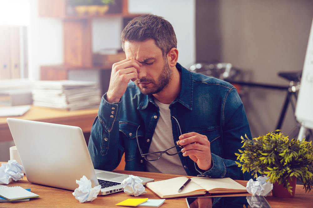 Ein müder junger Mann sitzt im Büro am Computer und leidet unter Stress.