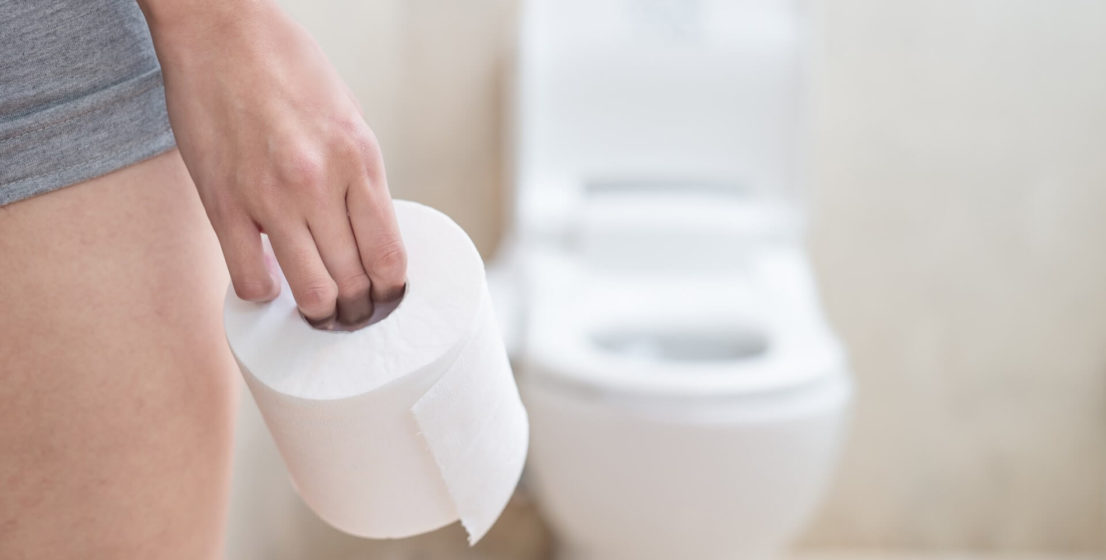 Eine Frau hält eine Rolle Toilettenpapier in der Hand und steht neben einer Toilettenschüssel.