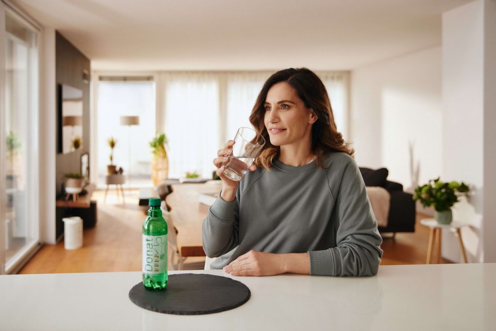 A woman sits at the kitchen counter and drinks a glass of Donat mineral water.