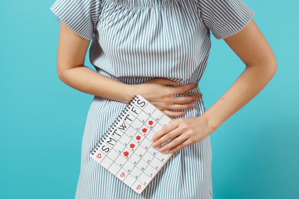 Женщина держится за живот, потому что во время менструации у нее вздутие живота, и она держит в руке календарь менструаций.