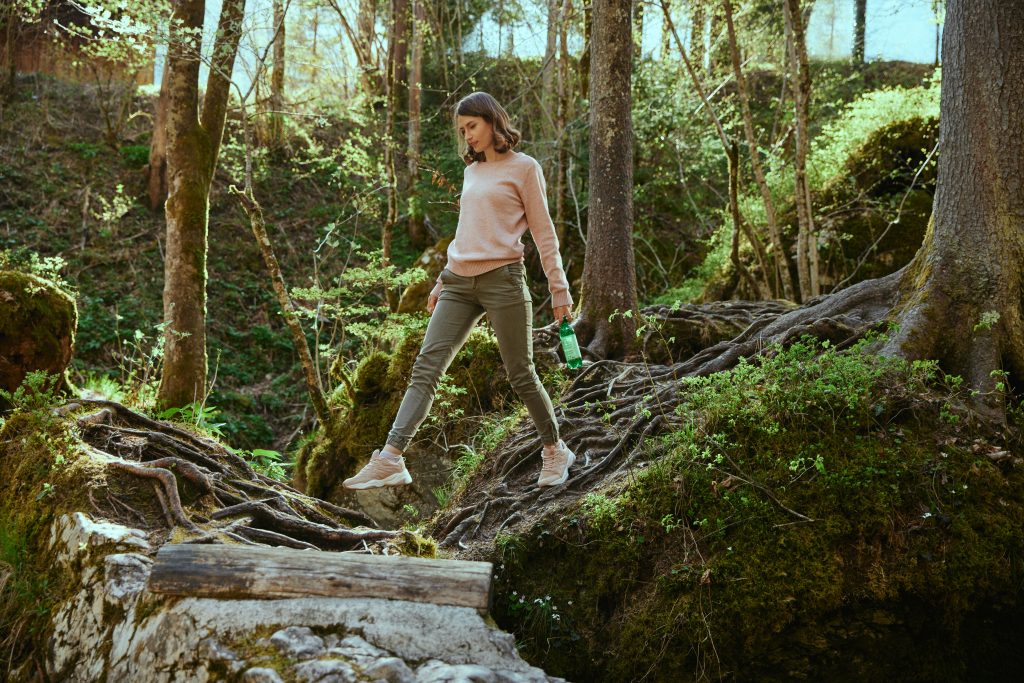 A woman jumps on the roots in the woods and holds a bottle of Donat in her hands.