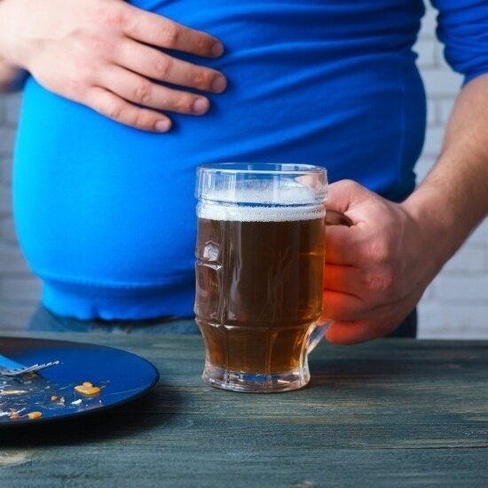 Ein Mann mit einem Krug Bier in der einen Hand, der sich mit der anderen am Bauch festhält.