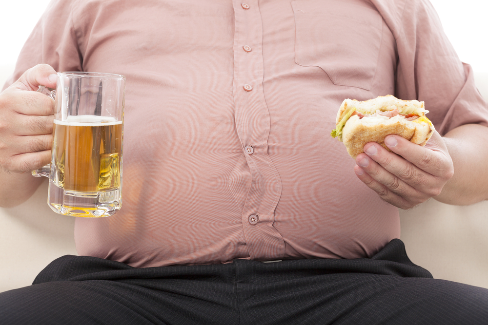 An overweight man is a hamburger and drinks beer