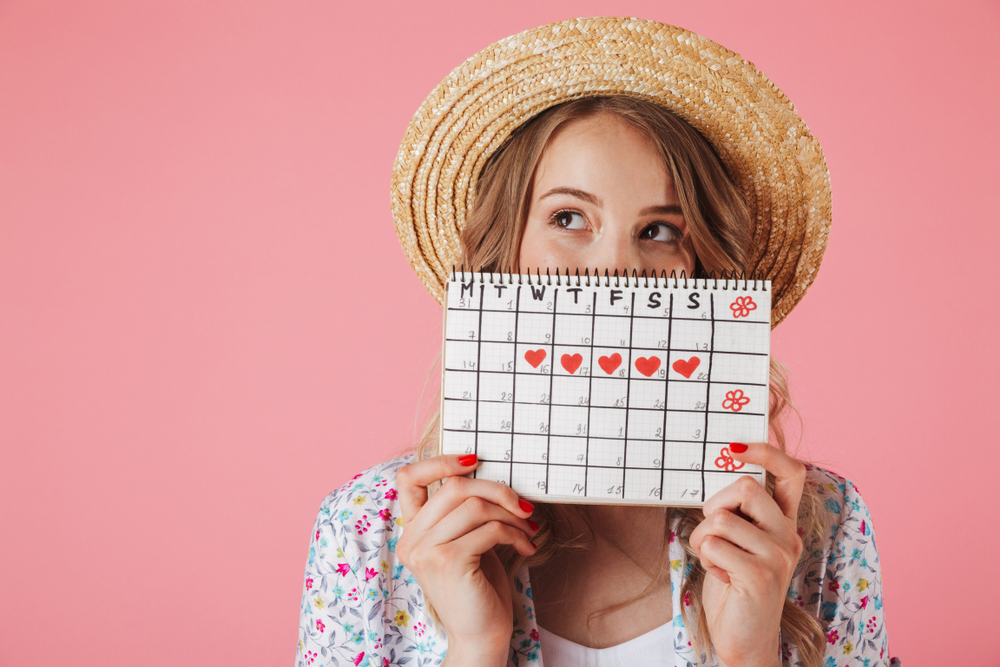 A young woman wearing a straw hat is holding a period calendar and is looking sideways – on a pink background.