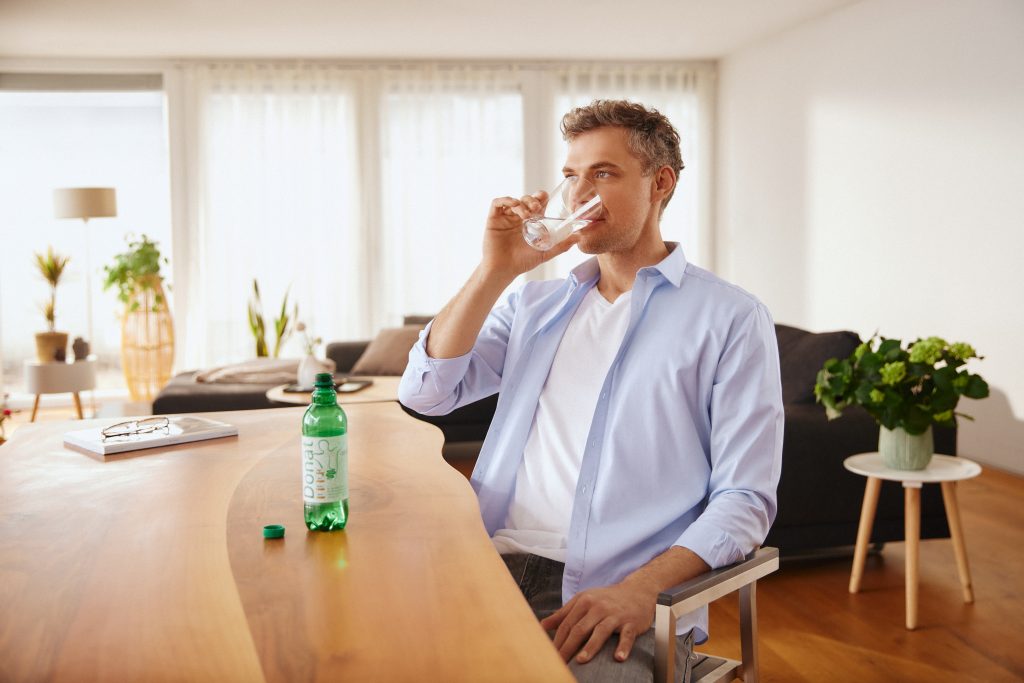 Мужчина сидит за деревянным обеденным столом и выпивает стакан свеженалитой минеральной воды «Донат».