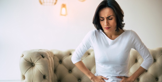 The woman in the table sits on the couch and holds her stomach with both hands as she is in pain.
