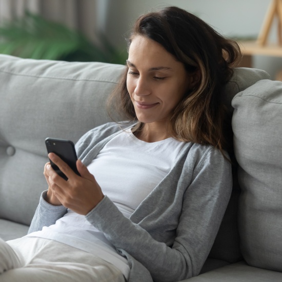 Eine lächelnde Frau ruht sich auf einer bequemen Couch im Wohnzimmer aus und surft auf einem modernen Smartphone im Internet.