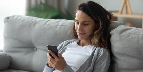 Nasmejana ženska počiva na udobnem kavču v dnevni sobi in brska po internetu na sodobnem pametnem telefonu.