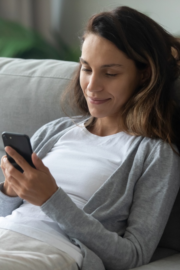 Una donna sorridente si riposa seduta confortevolmente sul divano in soggiorno e naviga in internet con uno smartphone.