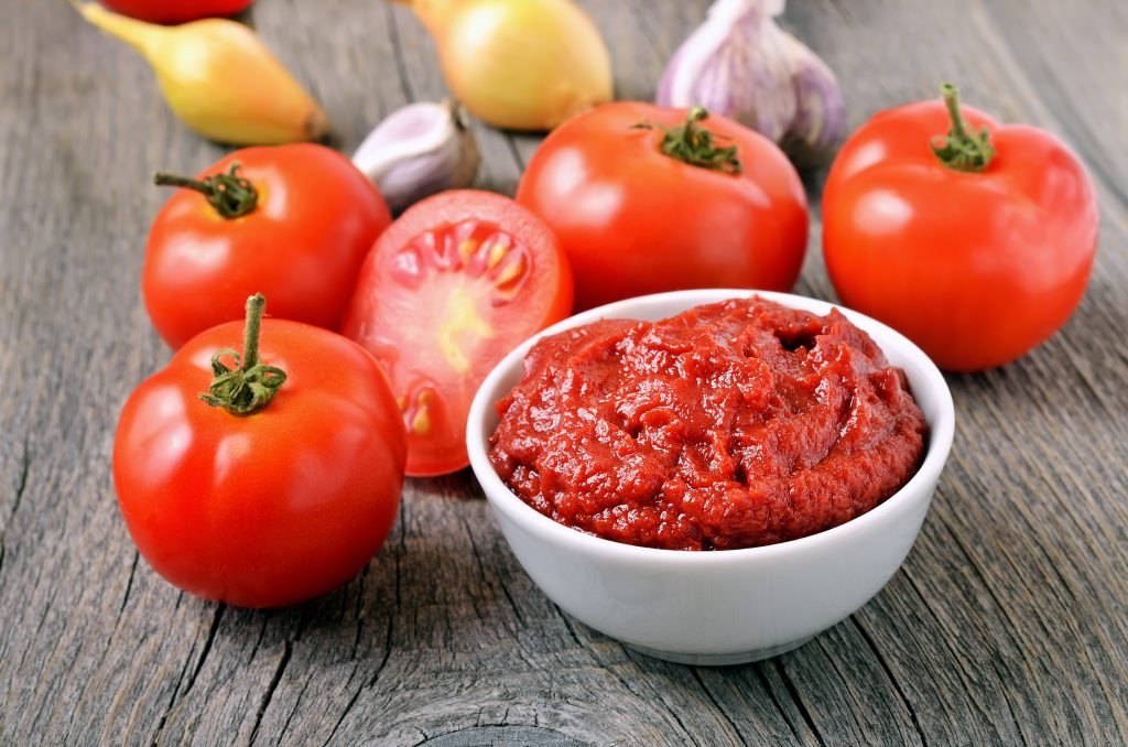 On a wooden table are tomatoes, onions, garlic and tomato sauce in a ceramic bowl.