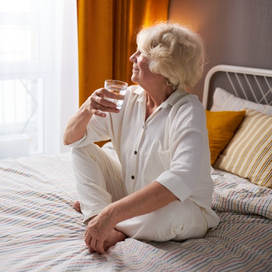 The older lady is sitting on the bed, drinking a glass of water and looking happily out the window.