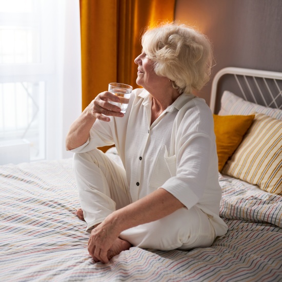 La signora più anziana è seduta sul letto, beve un bicchiere d'acqua e guarda felice fuori dalla finestra.