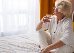 Die ältere Dame sitzt auf dem Bett, trinkt ein Glas Wasser und schaut glücklich aus dem Fenster.