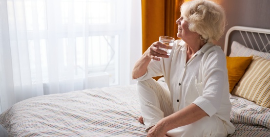 Die ältere Dame sitzt auf dem Bett, trinkt ein Glas Wasser und schaut glücklich aus dem Fenster.