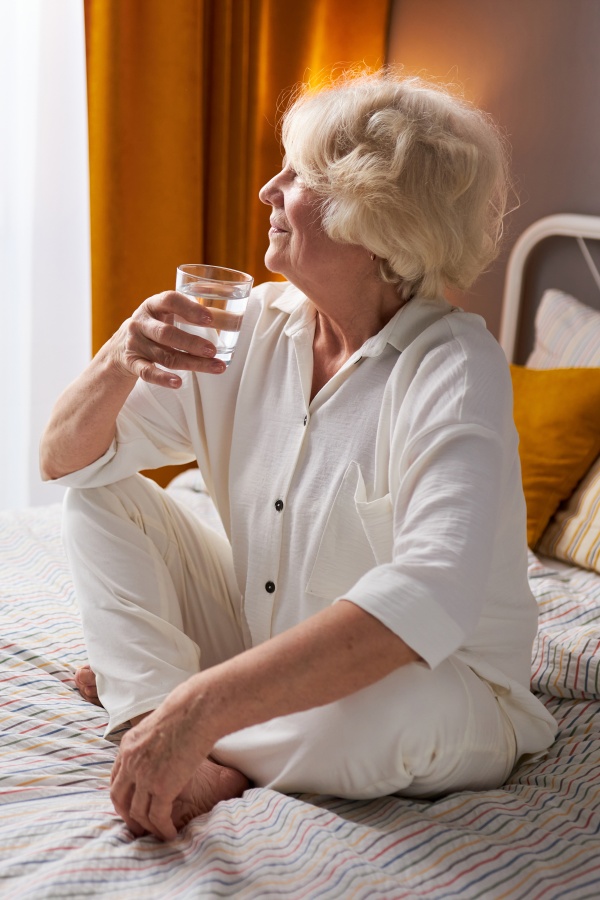 Starejša gospa v letih sedi na postelji, pije kozarec vode in veselo gleda skozi okno.